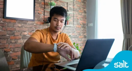 Man sitting at his home desk in front of his laptop looking at his watch.