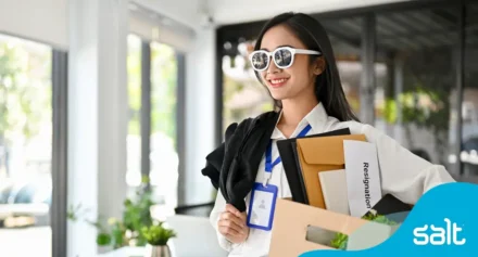 Woman wearing sunglasses, smiling and carrying a box with folders and a paper labeled “resignation”, symbolising a positive career transition.