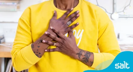 A person in a yellow sweatshirt, with tattooed arms, holds their hands over their heart in a warm, showing confidence