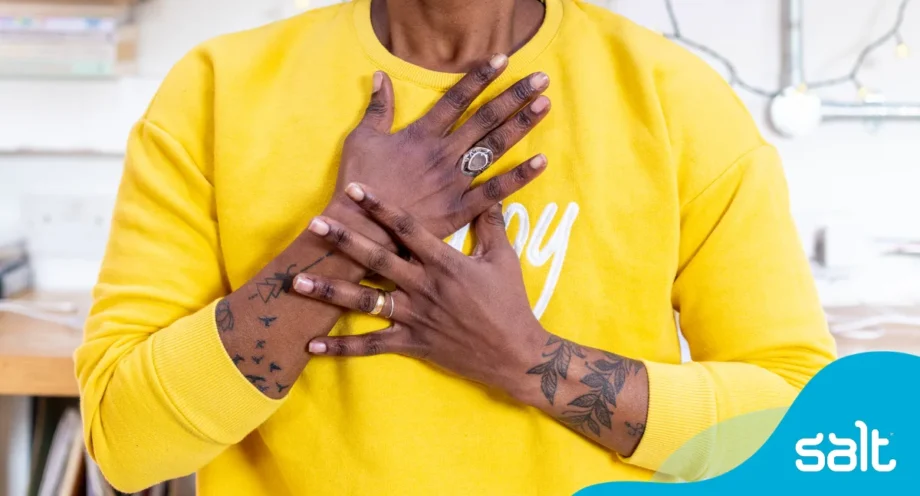 4 ways to be more confident at work and silence your inner critic 1 A person in a yellow sweatshirt, with tattooed arms, holds their hands over their heart in a warm, showing confidence