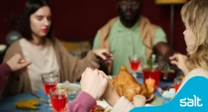 Diverse group of people holding hands around a Thanksgiving dinner table, symbolising unity and gratitude. Festive setting with a roast turkey, drinks, and decorations. Salt recruitment branding highlights connection and teamwork in ui/ux industry with a professional context.