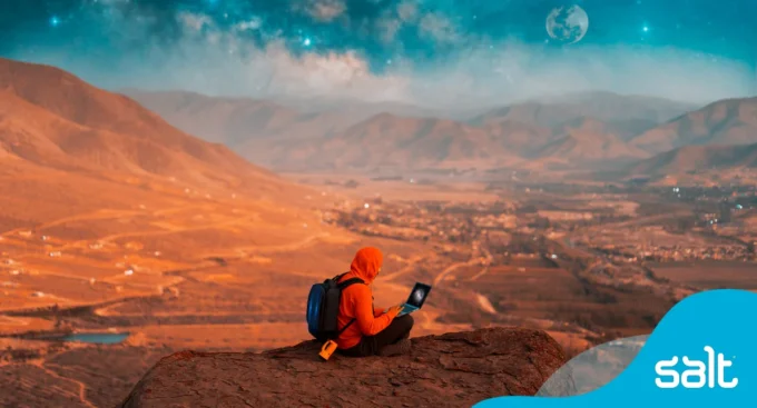 Man in red hoody sitting on-top of a mountain working on his laptop, symbolising a digital nomad