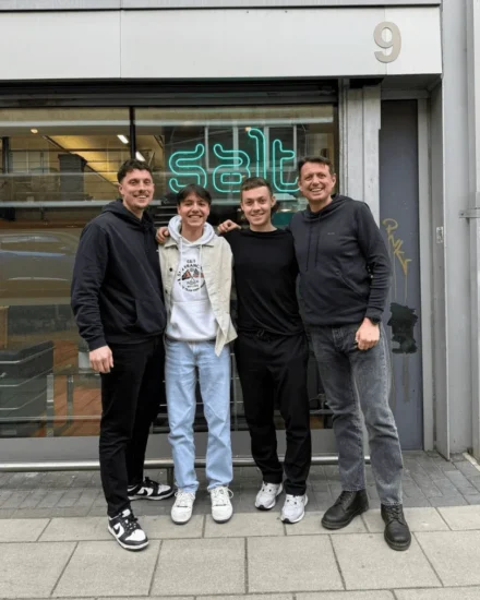 Four men smiling and posing together outside a building with a neon "Salt" sign