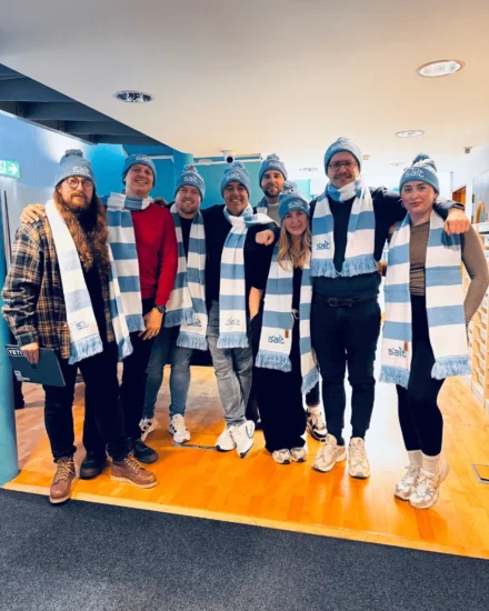 Group of eight people wearing matching blue and white "Salt" scarves and beanies, smiling indoors