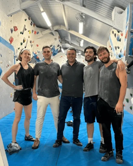 Group of five people smiling after indoor rock climbing, standing on a blue mat in a climbing gym