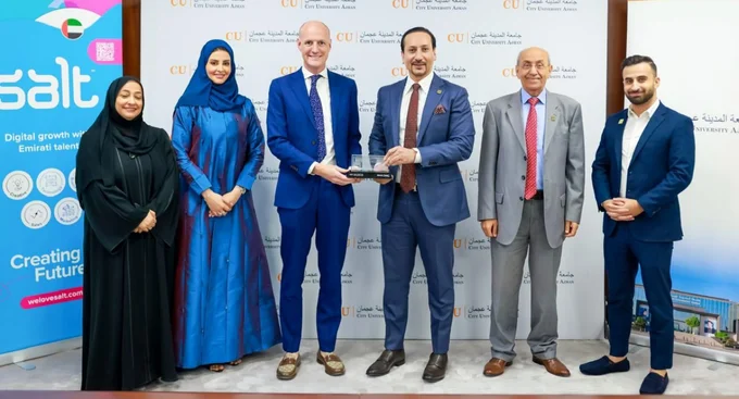 A group of six men and women in formal attire pose for a photo during a partnership event at City University Ajman, with branding from "Salt" in the background