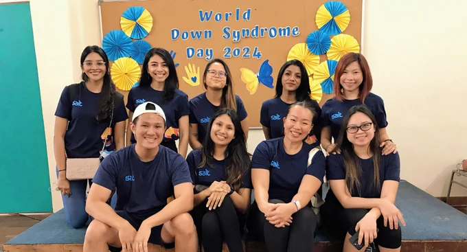A group of smiling volunteers in matching "salt" shirts pose together in front of a decorated board celebrating World Down Syndrome Day.