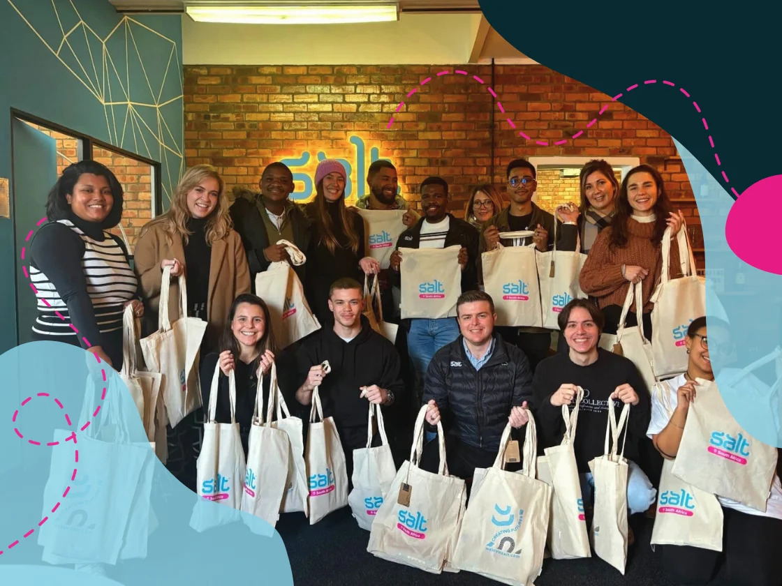 A group of people smiling and holding Salt-branded tote bags, gathered indoors for a charity initiative