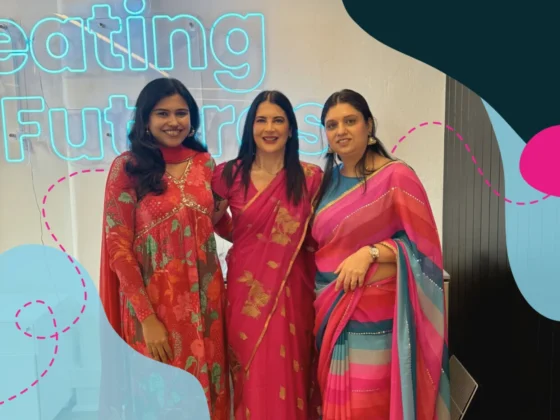 Three women dressed in vibrant traditional South Asian attire, smiling and standing together in front of a neon sign that reads "Creating Futures"