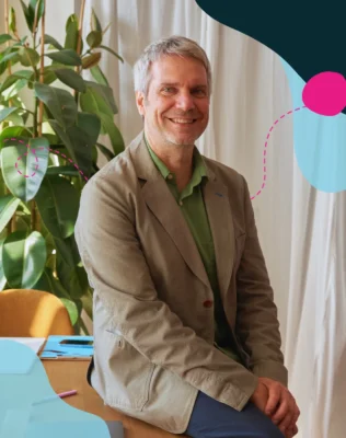 A middle aged man wearing a light brown blazer and green shirt sits on a table beside a large green plant smiling at the camera