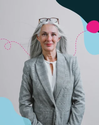 An elderly woman with long gray hair wears a grey blazer, stands confidently smiling at the camera