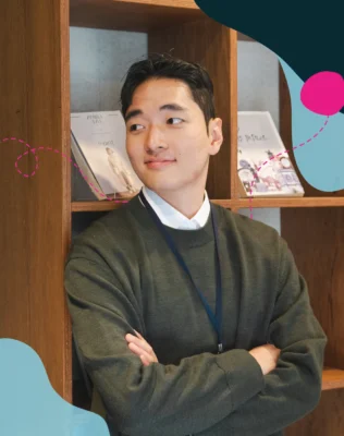 A young asian man stands confidently leaning with his back onto a book shelf armes crossed