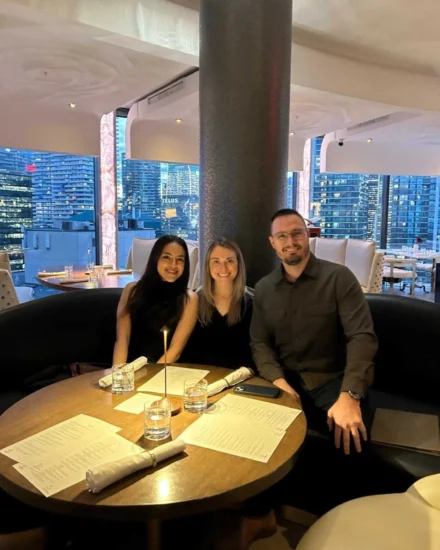 Three people smiling at a round dinner table in a modern restaurant with city skyline views through large windows