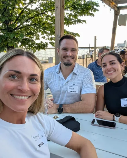 Three smiling people sitting at an outdoor table during a sunny day, wearing name tags for a tech event