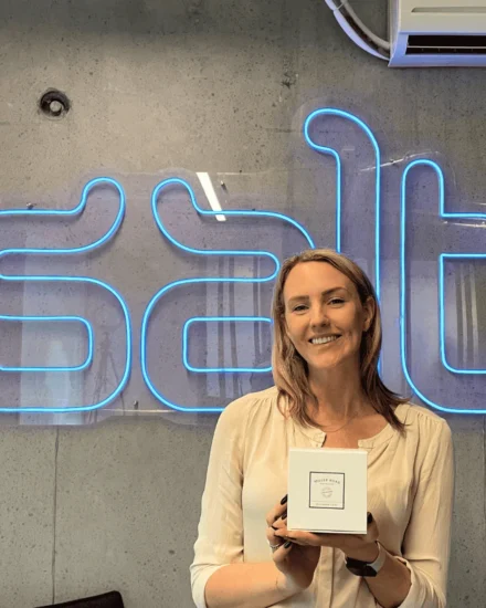 A woman smiling and holding a gift in her hands, while standing in front of a neon "Salt" sign