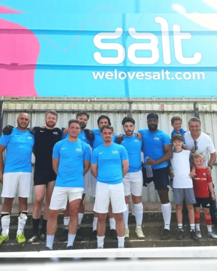 Football team in blue "Salt" jerseys posing together in front of a large "welovesalt.com" banner, joined by a few children
