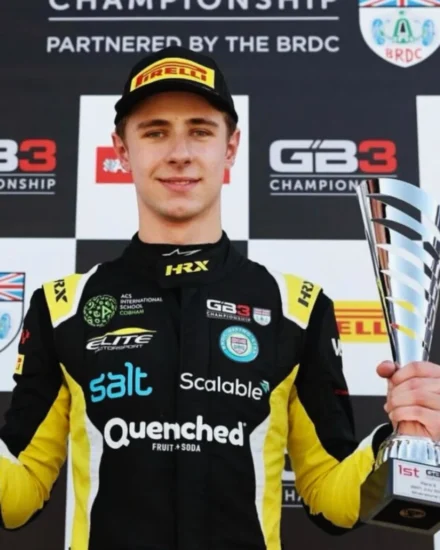 Race car driver, McKenzy Cresswell, holding a trophy on the GB3 Championship podium, wearing a black and yellow racing suit with sponsor logos, including Salt