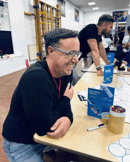 Man smiling while sitting at a table with educational brochures and colorful pens at a school event