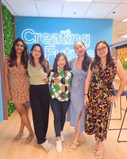 Five female colleagues posing and smiling in front of a "Creating Futures" neon sign