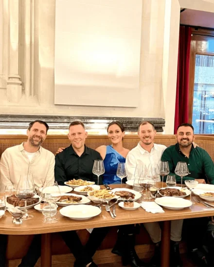 A group of six people smiling and seated at a restaurant table, enjoying a meal together