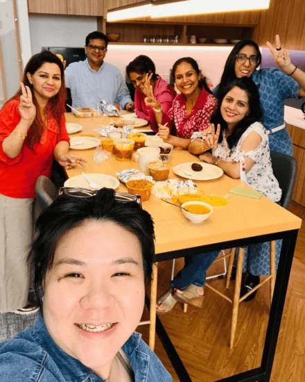 A group of colleagues enjoying a meal together around a table, smiling and posing with peace signs, dressed in colorful casual wear