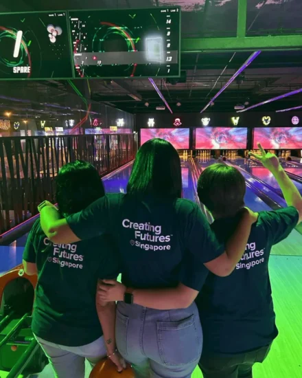 Three colleagues in matching "Creating Futures Singapore" shirts with arms around each other at a bowling alley