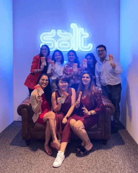 A group of colleagues smiling and holding festive envelopes in front of a neon "Salt" sign, dressed in red and white for a themed celebration