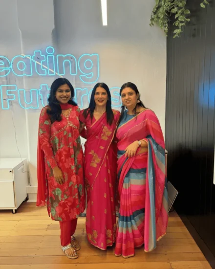 Three women dressed in vibrant traditional attire standing in front of a "Creating Futures" neon sign, smiling for the camera