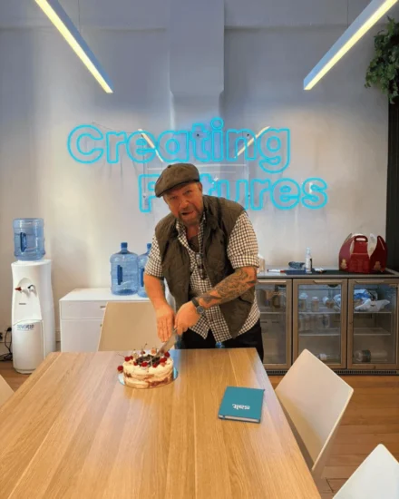 Man cutting a cake in an office kitchen with a "Creating Futures" neon sign behind him