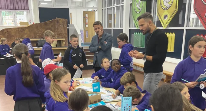 Group of schoolchildren in purple leavers hoodies sitting around tables with activities, guided by adults in a school hall.