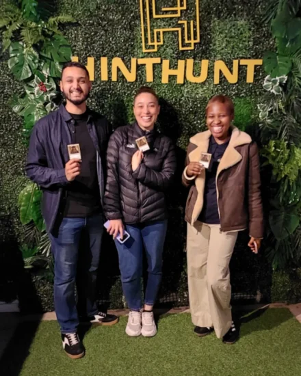 Three people smiling and holding cards in front of a HintHunt backdrop.