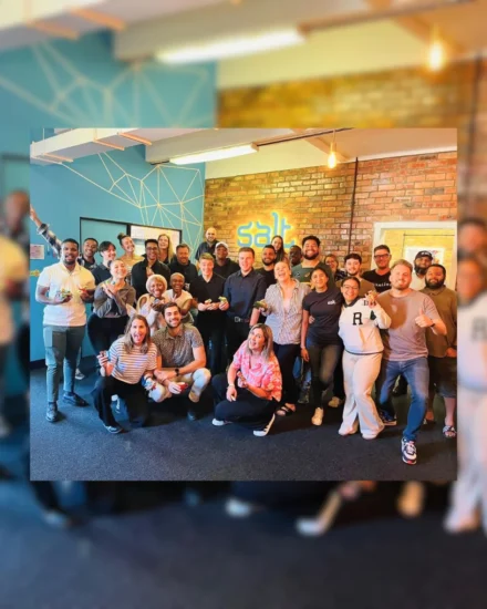 A large group of coworkers smiling and posing together in an office with a "Salt" sign on the wall, holding treats and celebrating.