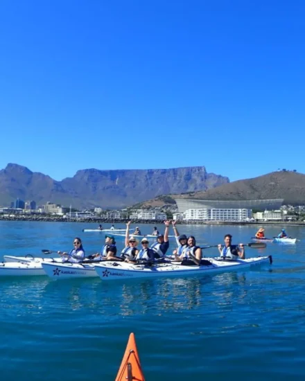A group of people in kayaks smiling and paddling on calm water with Table Mountain in the background on a clear, sunny day.