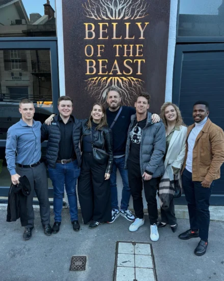 Group of seven people smiling in front of the Belly of the Beast restaurant sign.