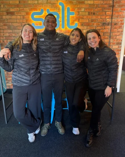 Four smiling team members wearing matching Salt-branded black jackets pose together in front of a brick wall with a glowing Salt logo.