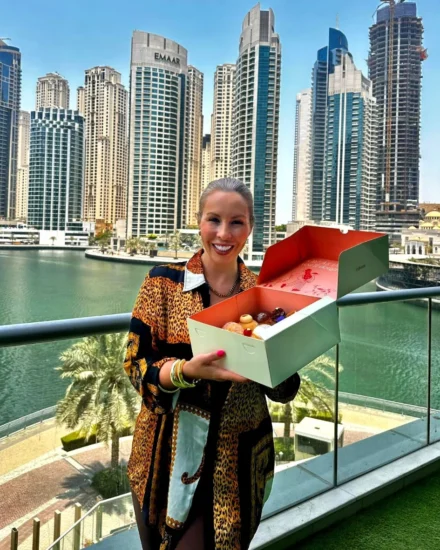 Smiling woman holding an open box of donuts on a balcony with the Dubai Marina skyline in the background.