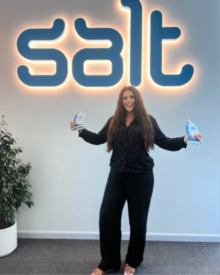 Smiling woman holding Salt awards in both hands, standing in front of a large illuminated Salt logo at the office.