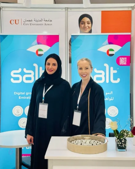 Two women standing at a Salt company booth during an event at City University Ajman, with branded banners and a display table in front of them.