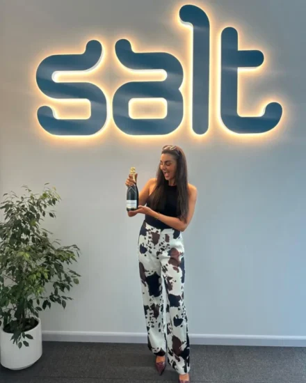 Smiling woman holding a champagne bottle in front of a glowing Salt logo at the office, celebrating a promotion.