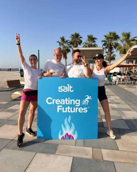 Four Salt team members posing cheerfully with a "Salt Creating Futures" sign at a beachside location, wearing activewear and making peace signs.