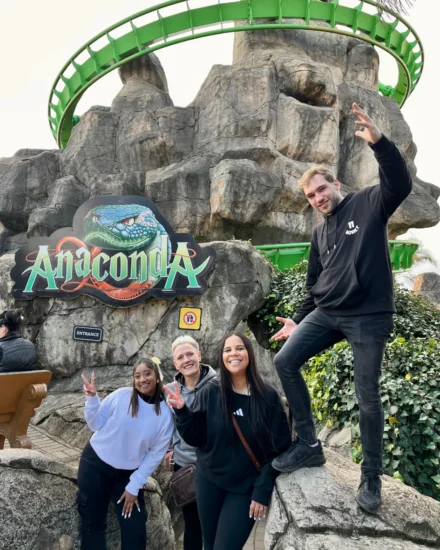 Four team members pose playfully in front of the Anaconda roller coaster entrance at Gold Reef City, smiling and making peace signs.