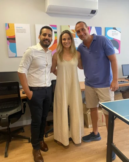 Three colleagues smiling and posing together in an office setting with colourful company value posters on the wall behind them