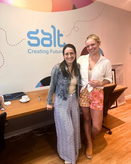 Two women smiling and standing in front of a "Salt Creating Futures" wall inside an office space.