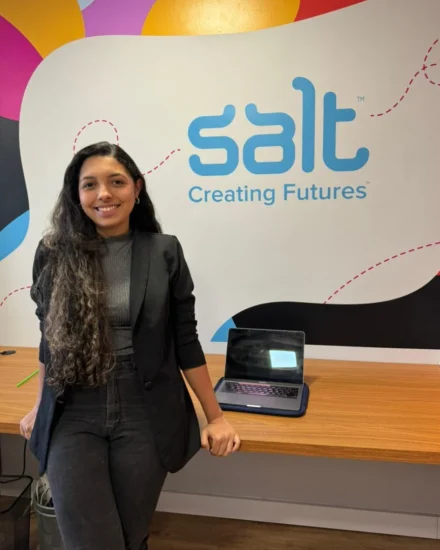 Smiling woman standing in front of a "Salt Creating Futures" wall inside an office, with a laptop on the desk beside her