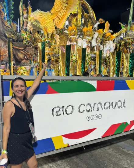 Smiling woman pointing at a vibrant float during Rio Carnival 2024, with colourful costumes and golden decorations in the background