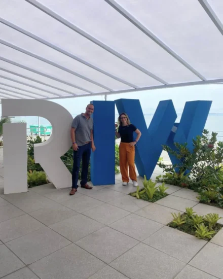 Two people posing in front of large "RIW" letters at Rio Innovation Week, with ocean and greenery in the background.