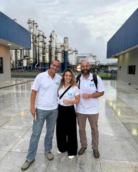Three people in matching white polo shirts standing outside an industrial plant, smiling at the camera during an agribusiness fair