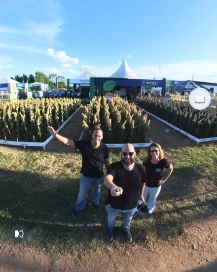 Three people smiling and posing for a selfie in front of maize crop displays at an outdoor agribusiness fair under a sunny blue sky
