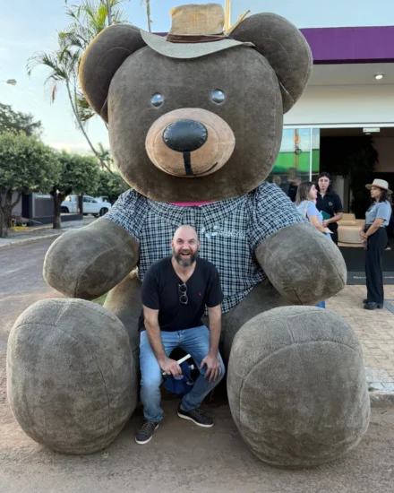Man smiling while sitting in front of a giant teddy bear statue wearing a hat and plaid shirt at the Agribusiness Fair.