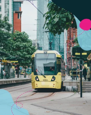 A yellow tram approaches a bustling city stop surrounded by trees, modern buildings, and waiting passengers on a cloudy day